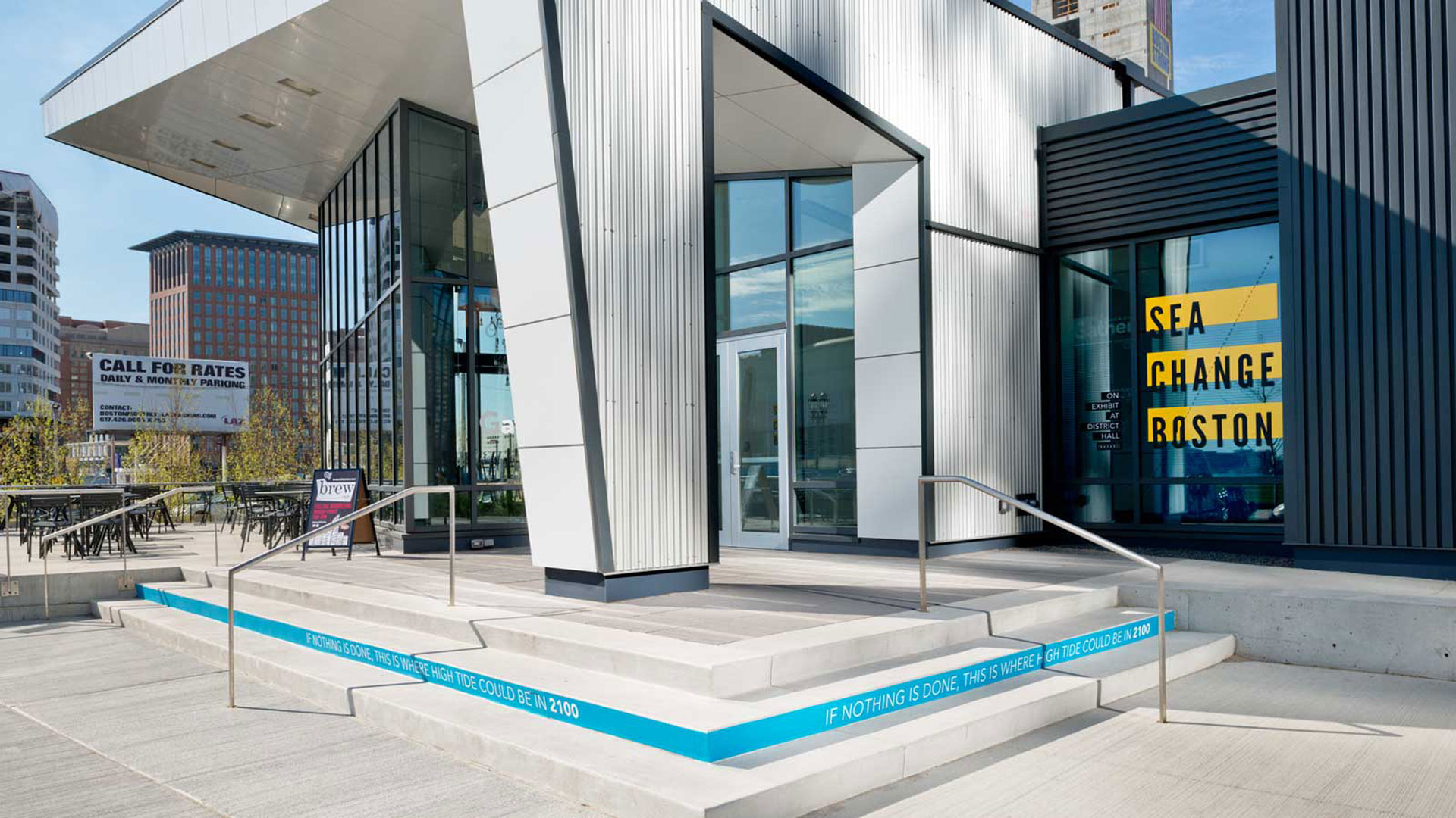 A photograph of the entrance of a modern building. There is a window on the right side with three yellow horizontal bars and typography. There is also a set of 3 steps in front of the building and one of the steps is blue with typography. Designs were made in Illustrator.